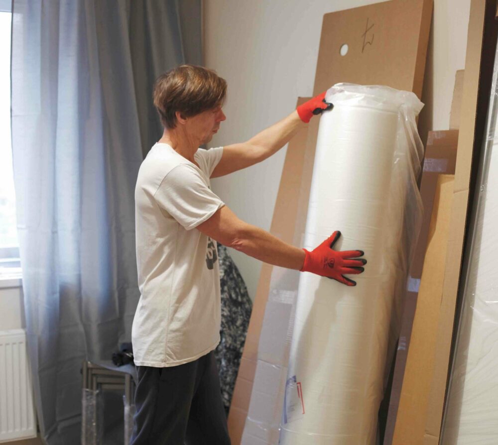 Umzugsunternehmen Hannover - Eine Person mit roten Handschuhen legt eine Rolle mit in Plastik eingewickeltem Material an eine Wand oder hebt sie auf. Sie befindet sich in einem Raum mit einem Fenster, blauen Vorhängen und verschiedenen anderen eingewickelten Gegenständen und Brettern, die an die Wand gelehnt sind, ähnlich einer Szene aus einem Umzugsunternehmen. Umzug Hannover