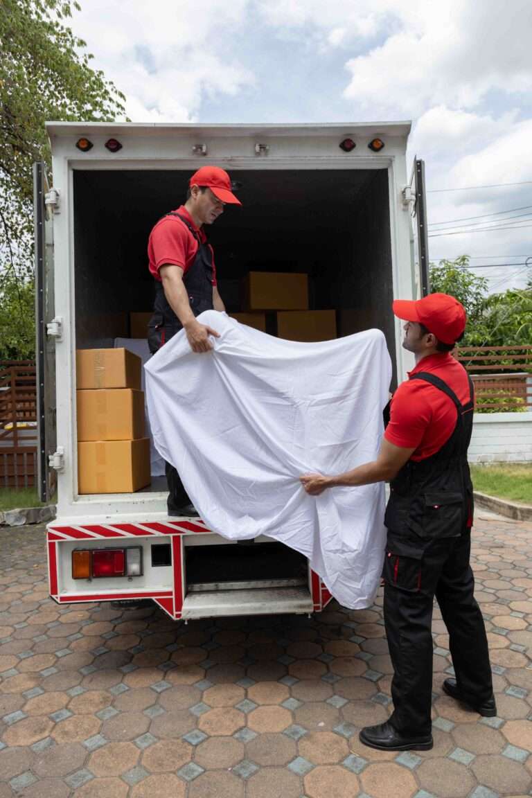 Umzugsunternehmen Hannover - Zwei Lieferarbeiter in roten Hemden, Mützen und schwarzen Overalls von einem Umzugsunternehmen stehen neben einem offenen Lieferwagen. Ein Arbeiter steht auf dem Lastwagen und hält ein weißes Laken, während der andere draußen steht und das andere Ende festhält. Der Lastwagen enthält mehrere braune Kartons. Umzug Hannover