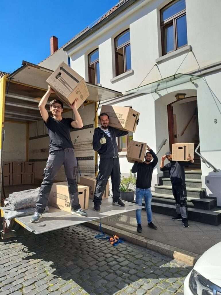 Umzugsunternehmen Hannover - Vier Personen, leger gekleidet, stehen auf der Ladefläche eines Umzugswagens vor einem Gebäude. Sie lächeln und halten Kisten hoch. Es sieht so aus, als würden sie gerade umziehen oder ausladen. Das Gebäude hat weiße Wände und große Fenster. Umzug Hannover