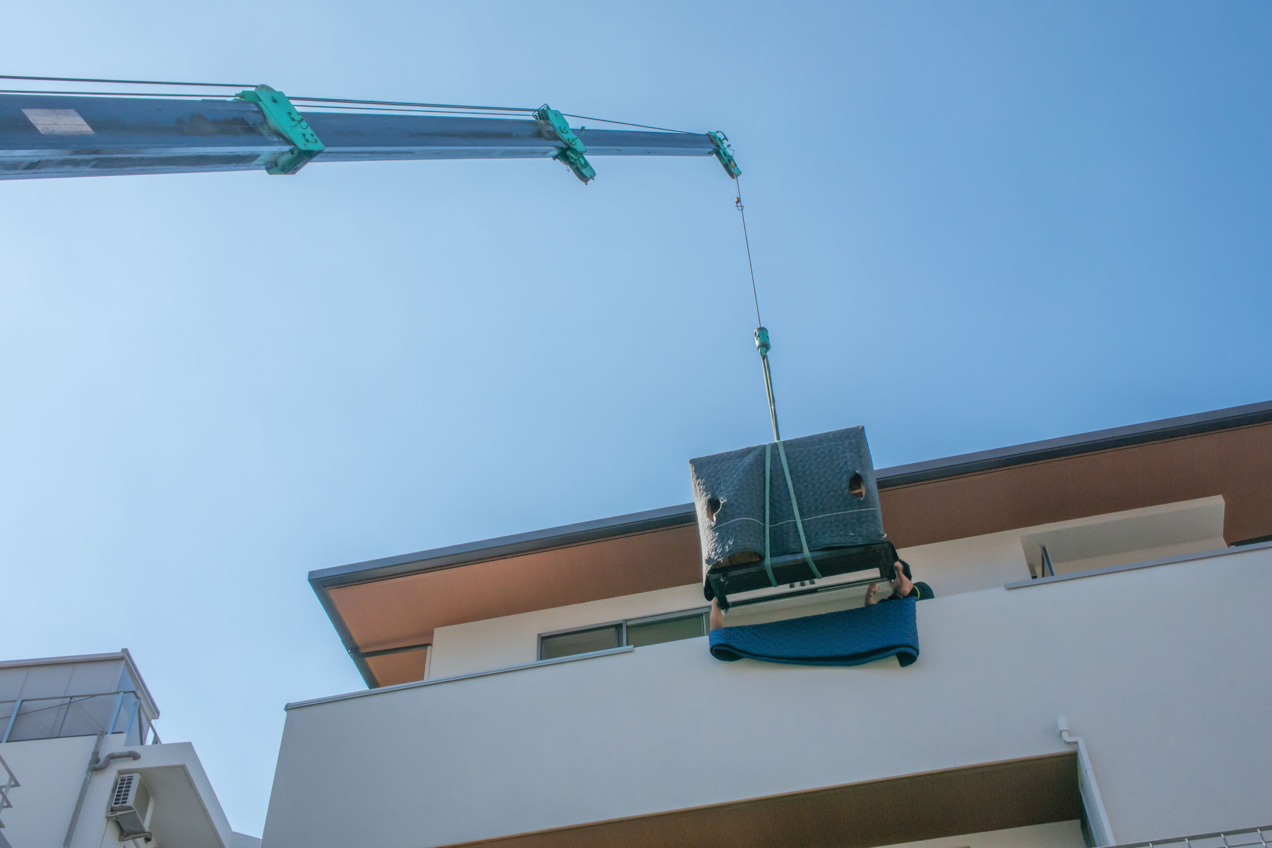 Umzugsunternehmen Hannover - Ein Kran hebt ein großes, verpacktes Objekt vom Boden auf den Balkon einer Wohnung im oberen Stockwerk eines weißen Gebäudes, das an einen filigranen Klaviertransport erinnert. Der klare blaue Himmel umrahmt die Szene, wobei ein Teil des Kranarms und des Kabels nach oben ragt. Umzugsunternehmen Seelze.