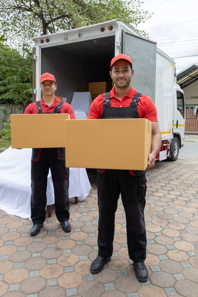 Umzugsunternehmen Hannover - Zwei Lieferarbeiter in roten Mützen und Hemden halten Kartons neben einem offenen Lastwagen des Umzugsunternehmens Garbsen. Der Lastwagen steht auf einer gepflasterten Fläche mit Bäumen und einem Gebäude im Hintergrund. Umzugsunternehmen Seelze.