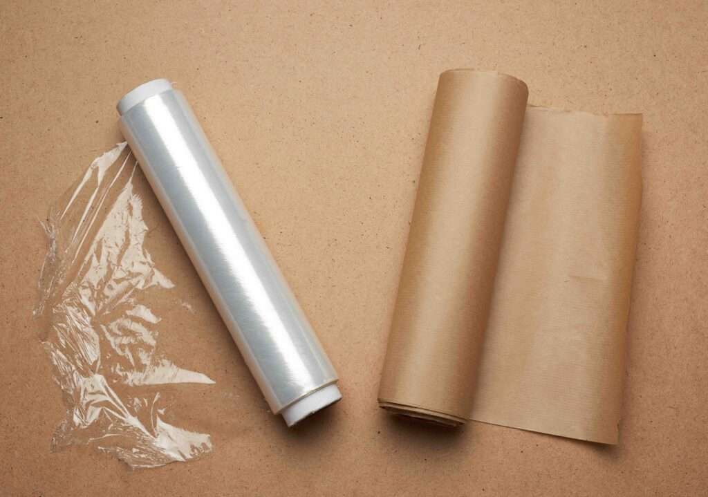 Umzugskartons 21 Umzugsunternehmen Hannover - Eine Rolle Klarsichtfolie und eine Rolle braune Papierfolie liegen nebeneinander auf einer braunen Fläche, links ist noch etwas Folie ausgerollt – ideal, wenn Sie Strechfolie in Hannover kaufen möchten. Umzugsunternehmen Seelze.
