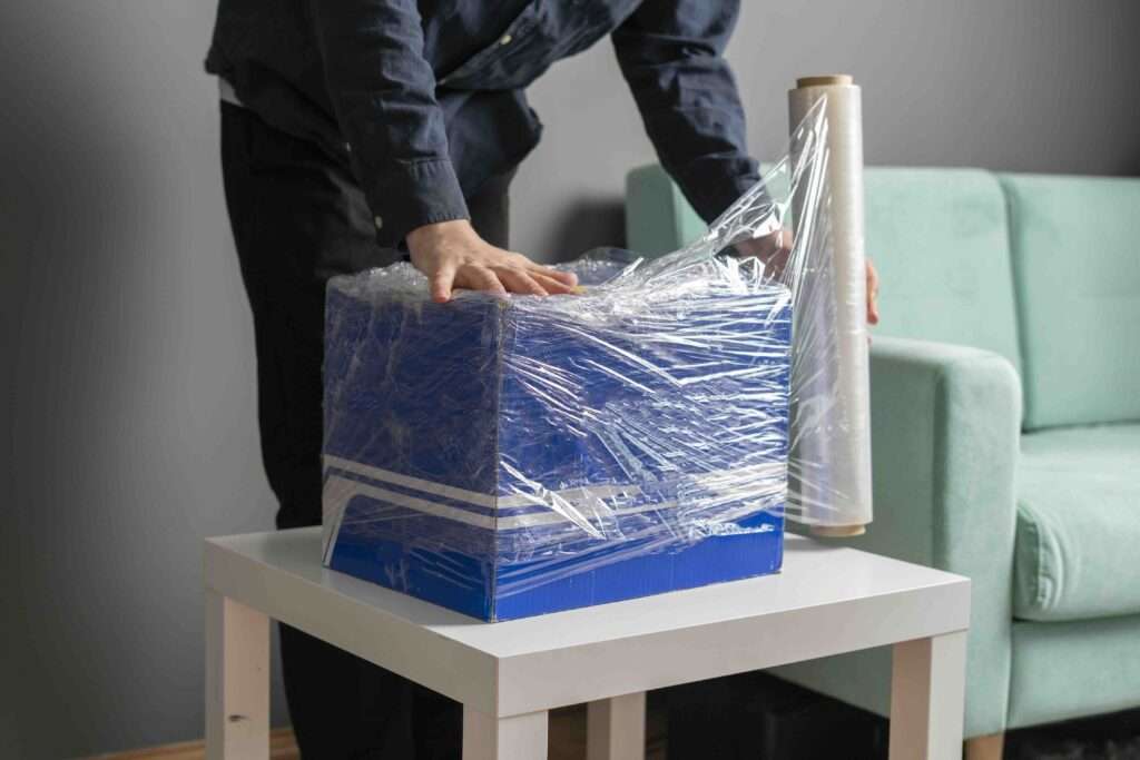 Umzugsunternehmen Hannover - Eine Person verpackt einen blauen Karton mit durchsichtiger Plastikfolie auf einem weißen Tisch. Sie verwendet eine Rolle Plastikfolie – ideal für alle, die Strechfolie in Hannover kaufen möchten. Im Hintergrund ist ein hellgrünes Sofa zu sehen. Umzugsunternehmen Seelze.