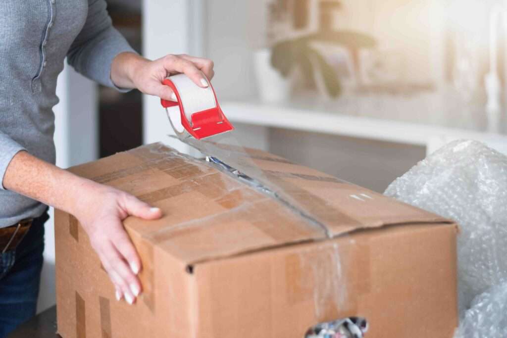 Umzugskartons 9 Umzugsunternehmen Hannover - Eine Person verschließt einen Karton mithilfe eines Klebebandabrollers mit durchsichtigem Klebeband; Luftpolsterfolie daneben deutet darauf hin, dass Gegenstände verpackt oder transportiert werden müssen – perfekt für alle, die Verpackungsmaterial für den Heim- oder Bürogebrauch in Hannover kaufen möchten. Umzugsunternehmen Seelze.