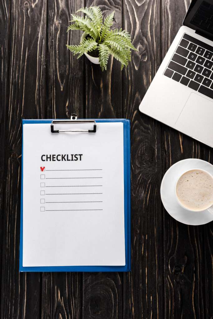 Checkliste Umzug 1 Umzugsunternehmen Hannover - Auf einem dunklen Holzschreibtisch liegt neben einem Laptop, einer kleinen Topfpflanze und einer Tasse Kaffee ein Klemmbrett mit einer Umzugs-Checkliste (ein Kästchen ist abgehakt). Umzugsunternehmen Seelze.